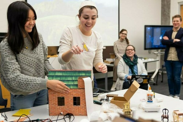Smiling students working with wires, glue, and a dollhouse-shaped printer in a classroom while professors watch