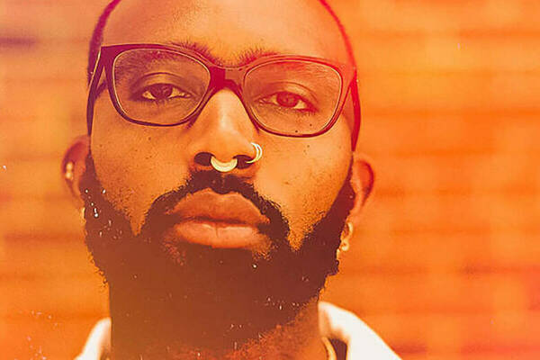 Joseph Earl Thomas, presenting as a black male with a beard, glasses, and nose piercings. He is standing in front of a brick wall and the photo has a orange-sepia filter added.