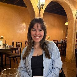 Smiling young woman with brown hair in a light blue shirt in a restaurant with warm orange walls and arches.