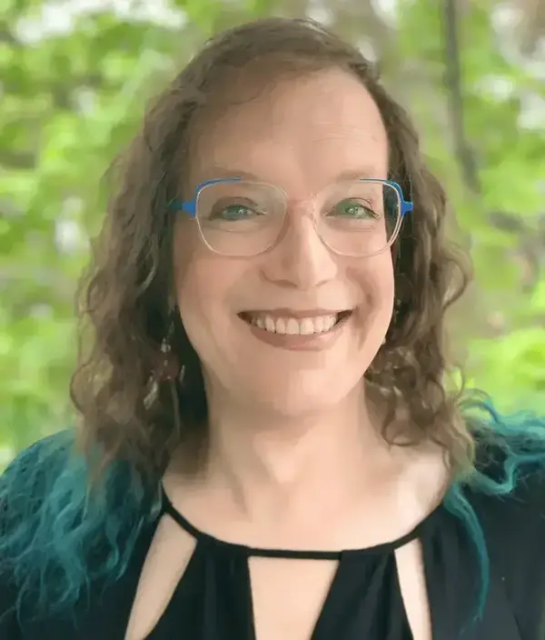 Smiling woman with wavy brown hair with teal tips, wearing blue-accented glasses and a black cutout top.